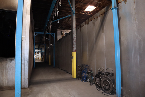 Industrial corridor in a factory with blue steel beams, overhead piping, and hose reels along the right wall.