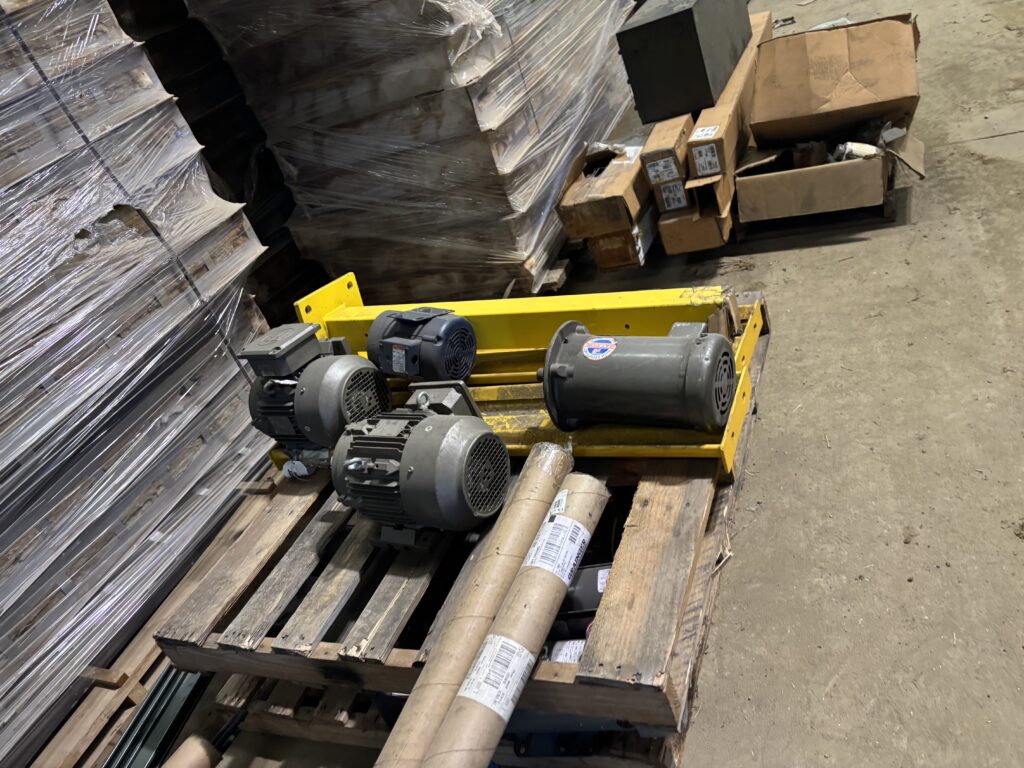 Pallets stacked with wrapped bundles and several industrial motors resting on a yellow pallet jack in a warehouse.