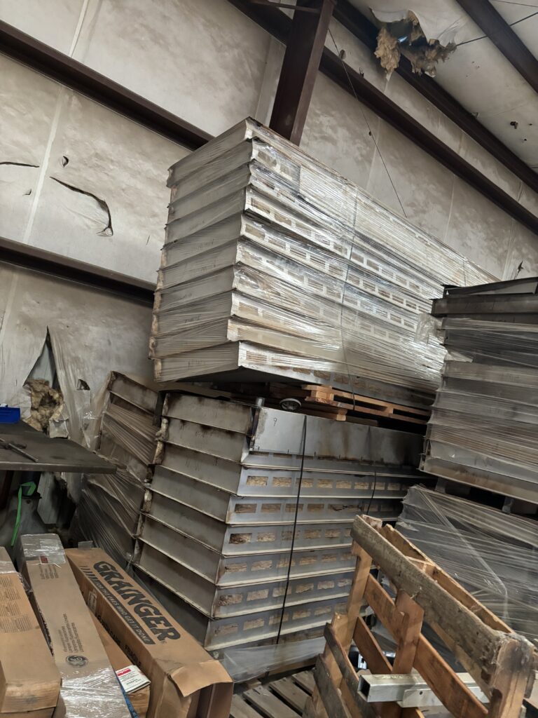 Stacks of wrapped metal panels on pallets in a dusty, cluttered warehouse. A damaged wall and exposed ceiling are visible in the background.