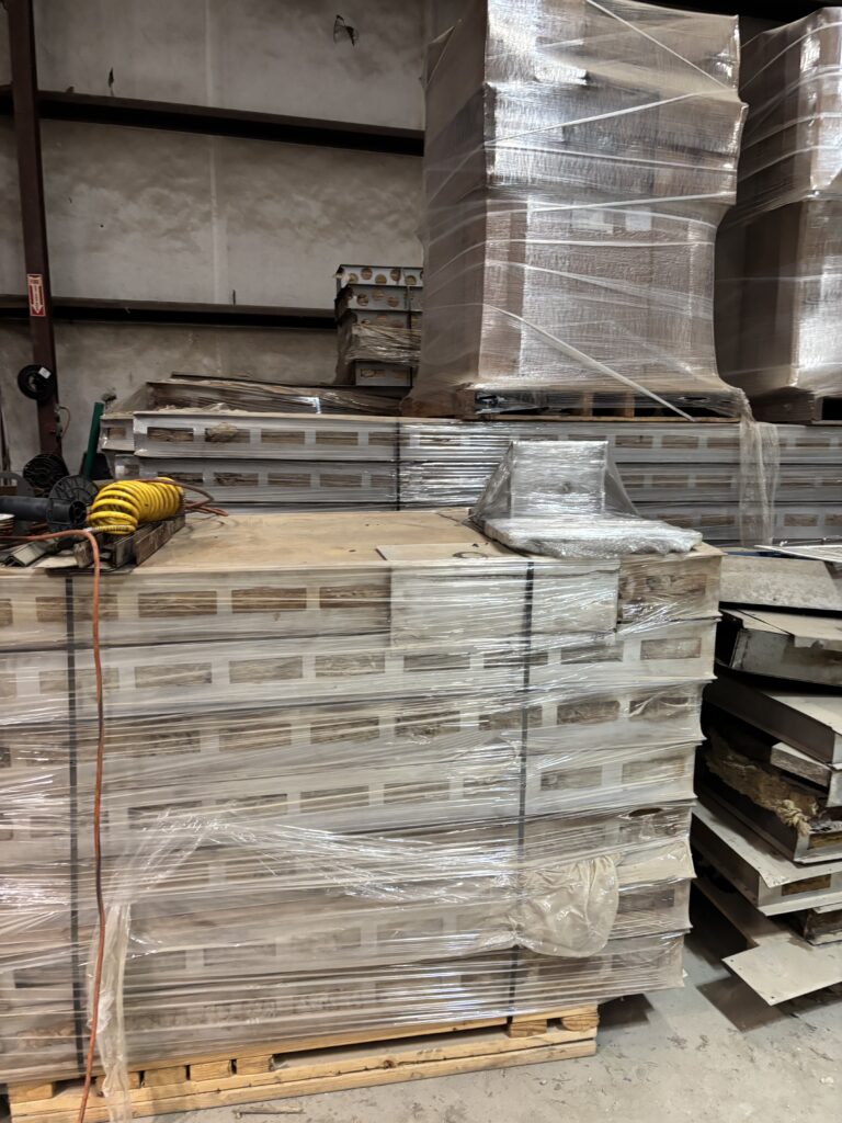 Warehouse scene with stacked pallets wrapped in plastic, containing metal panels or boards, in a storage area with a coiled yellow hose on the floor.