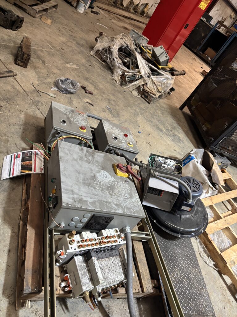 Industrial workshop floor with scattered electrical panels, wires, and machinery amidst debris and pallets; red pillar in the background.