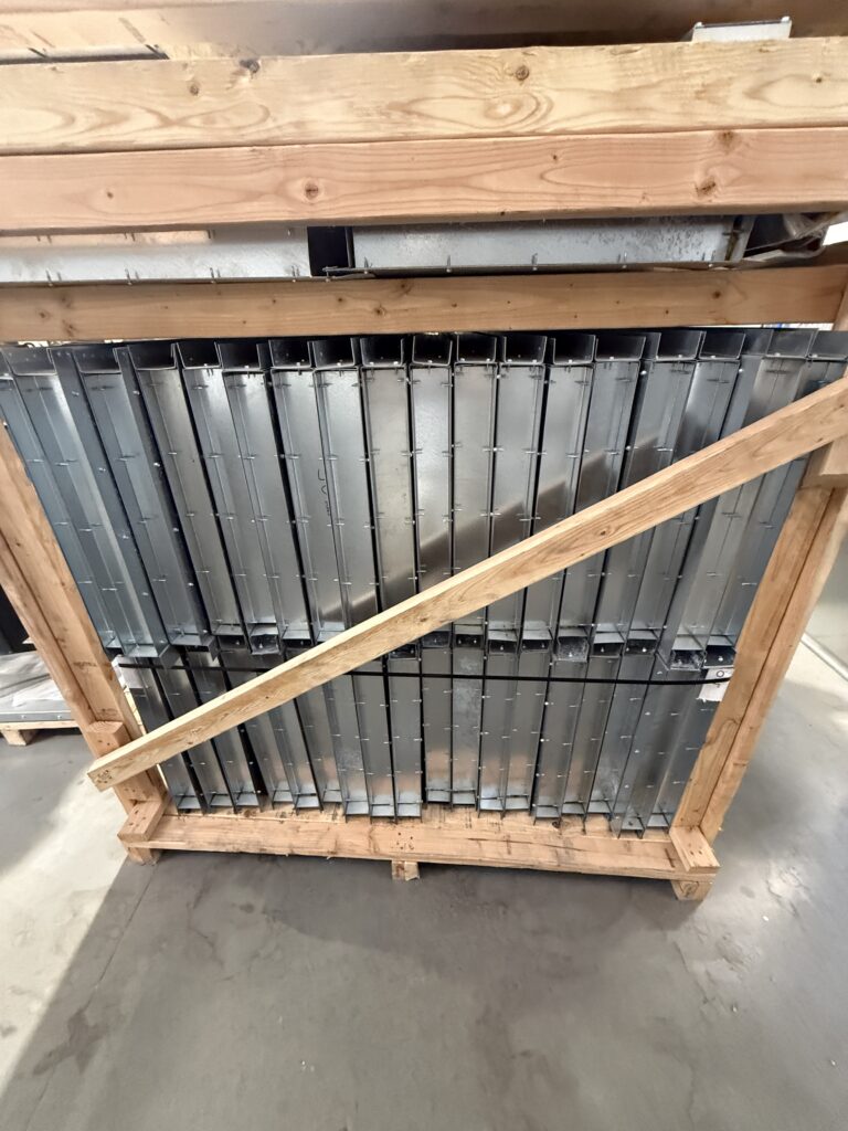 Stack of galvanized metal louvered panels secured in a wooden crate, likely HVAC duct components ready for transport