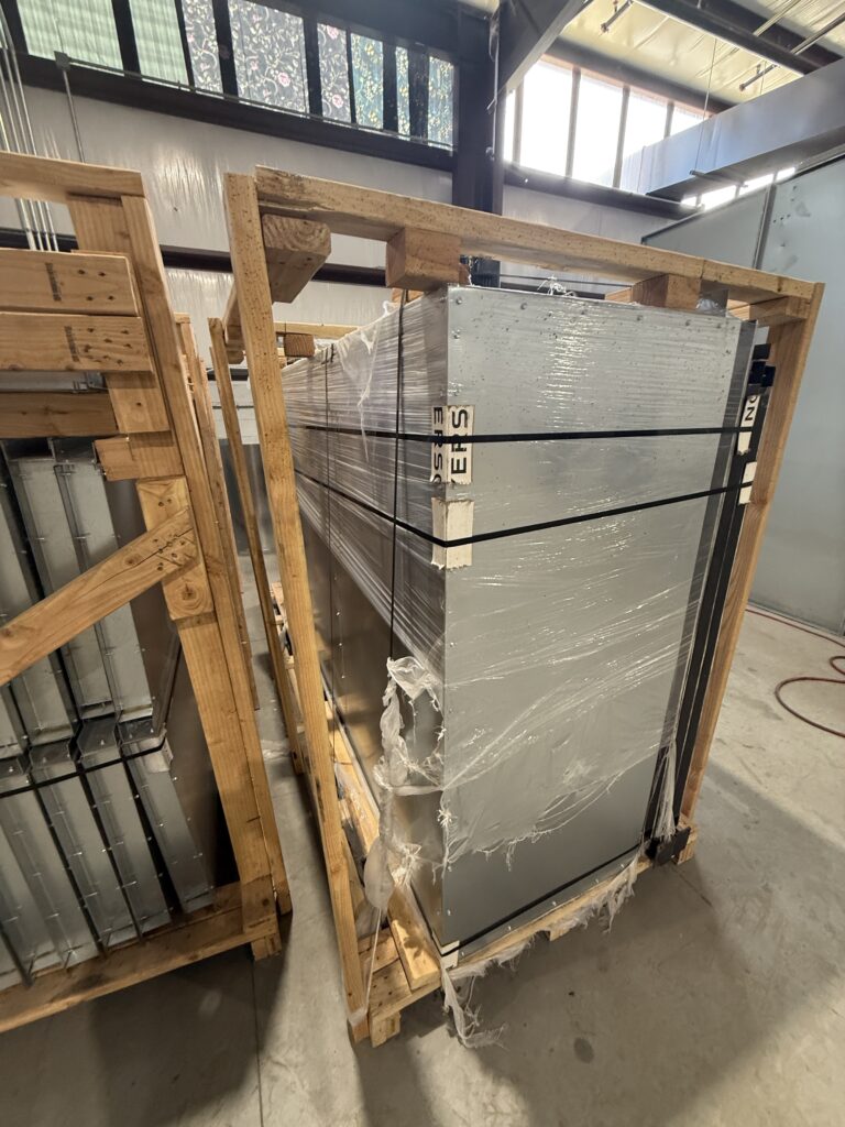 Large industrial metal equipment wrapped in plastic, strapped to a wooden pallet, in a warehouse with concrete floor and overhead beams.