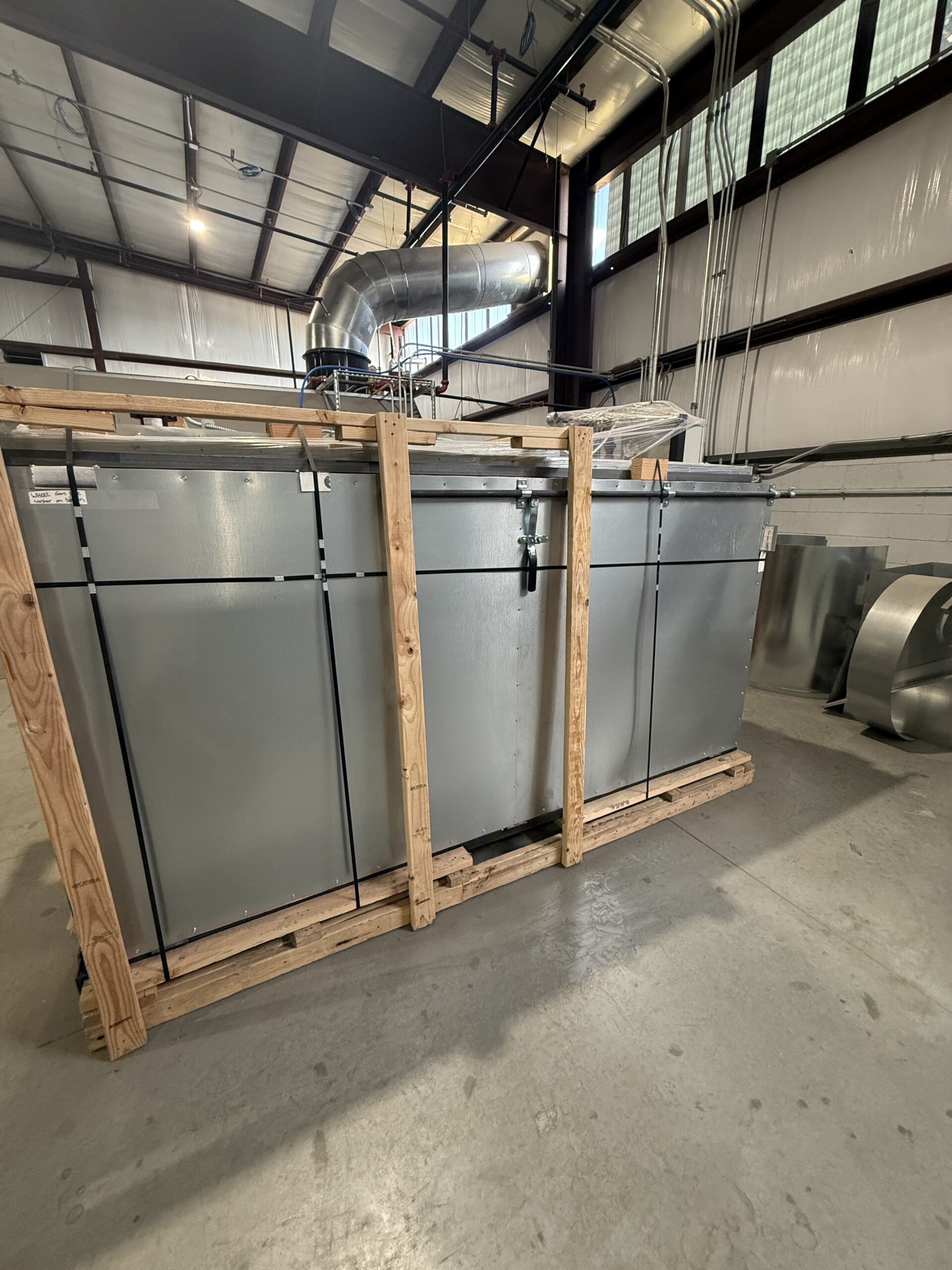 Large gray industrial HVAC unit on a wooden pallet frame inside a warehouse, with overhead ducts, cables, and metal framing visible.
