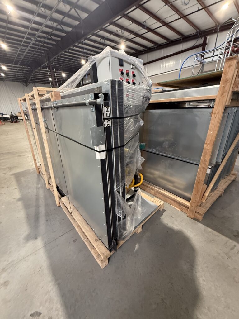 Large industrial metal cabinet with a control panel, wrapped in plastic and strapped to a wooden crate in a warehouse setting.