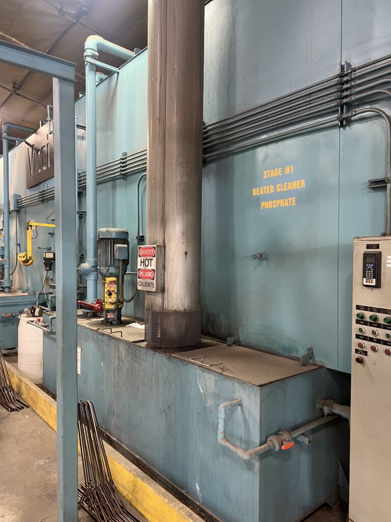 Industrial processing equipment: a large blue machine with multiple pipes and a control panel, plus warning signs reading 'Stage #1 Heated Cleaner Phosphate' and 'Danger Hot'