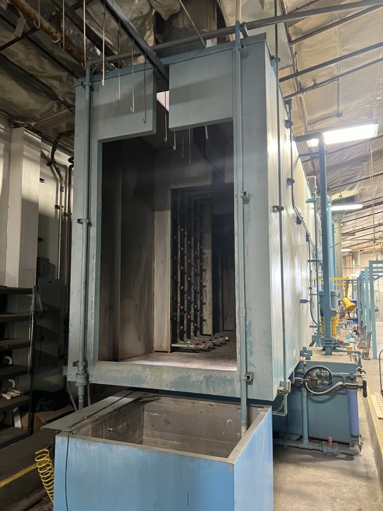 Blue industrial cabinet or oven housing in a factory with hanging hooks above and pipes along the ceiling, suggesting a processing area.
